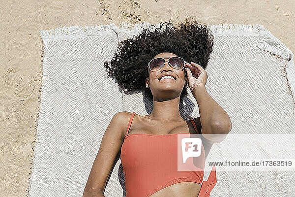Lächelnde junge Frau mit lockigem Haar und Sonnenbrille  die auf einem Strandtuch liegt