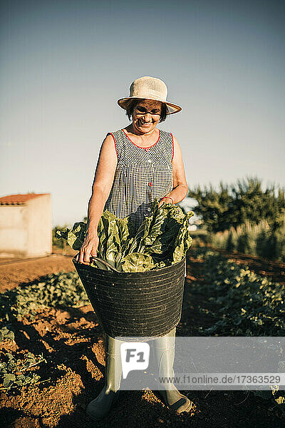 Ältere Bäuerin lächelt  während sie einen Korb im Gemüsegarten hält