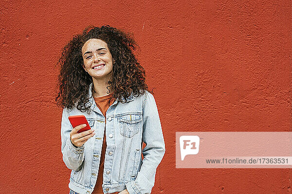 Lächelnde Frau hält ihr Smartphone vor einer roten Wand