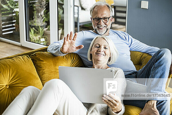 Smiling man waving hand during video call through laptop while sitting with woman on sofa