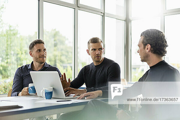 Male professionals sitting with laptop while discussing at desk in office