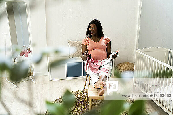 Glückliche schwangere Frau schaut weg  während sie mit Babykleidung auf einem Stuhl im Schlafzimmer sitzt