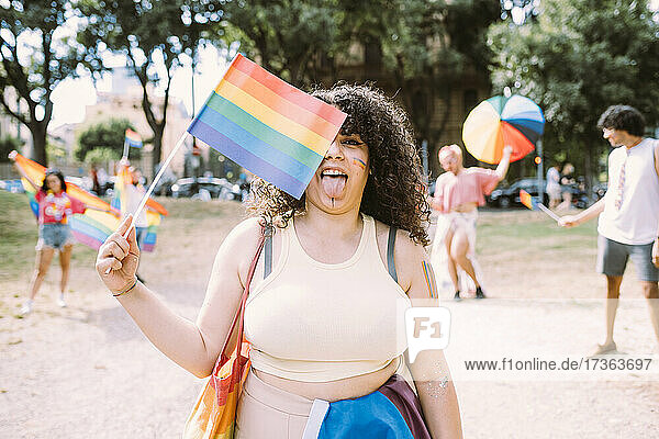 Junge Frau hält Regenbogenflagge  während Freunde im Hintergrund im Park