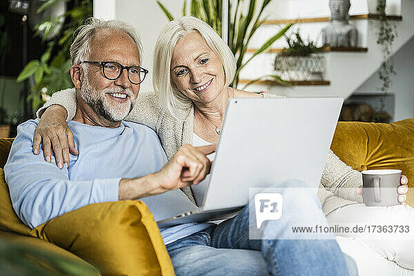 Smiling mature man using laptop while sitting with woman on sofa at home
