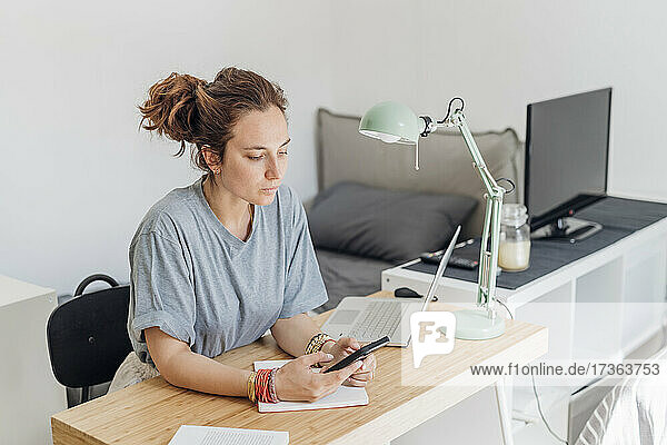 Female freelancer using mobile phone while sitting with laptop at apartment
