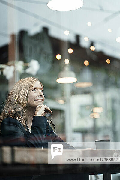 Thoughtful female professional sitting with hand on chin in cafe seen through glass window