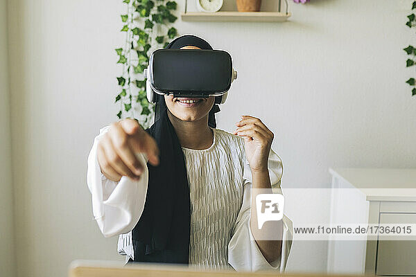 Smiling female freelance worker pointing while using virtual reality headset at home