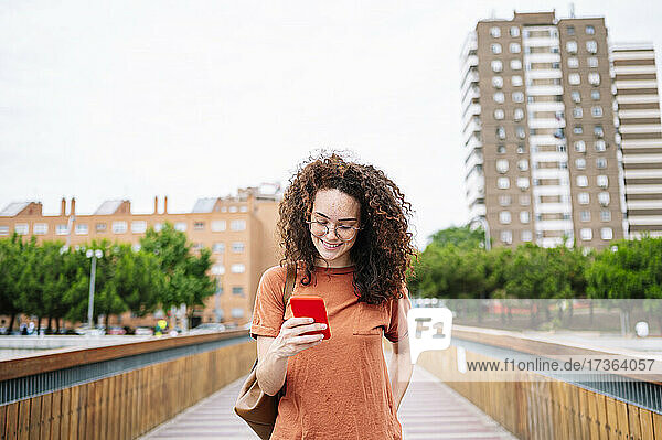 Lächelnde  lockig behaarte Frau  die ihr Smartphone benutzt  während sie auf einer Brücke spazieren geht