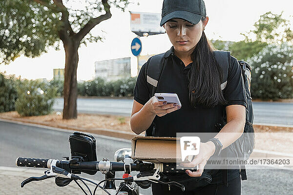 Junge Zustellerin mit Fahrrad  die ein Paket über ihr Smartphone scannt