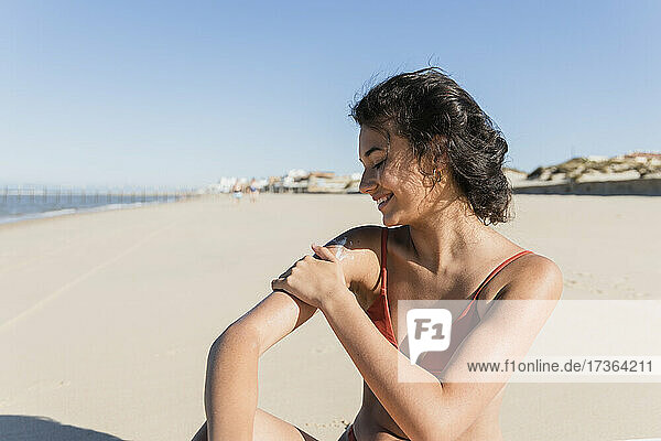 Lächelnde junge Frau  die Sonnencreme aufträgt  während sie an einem sonnigen Tag am Strand sitzt