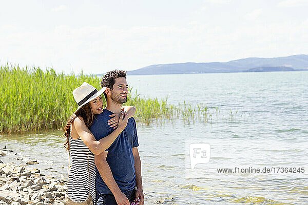 Smiling woman with hat embracing man from behind while standing by Lake Trasimeno