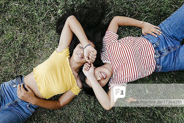 Smiling lesbian couple covering eyes with hands while lying on grass at park