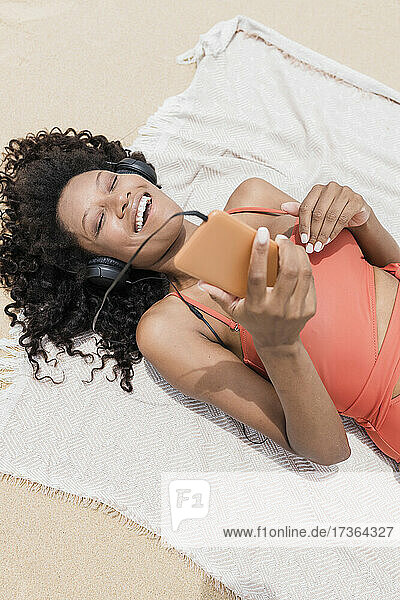 Fröhliche Frau mit geschlossenen Augen hört Musik  während sie auf einem Strandtuch liegt