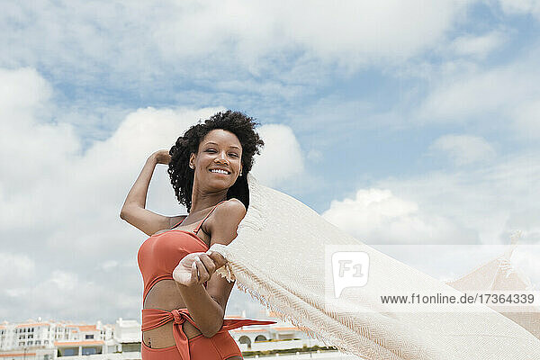 Lächelnde schöne junge Frau hält Strandtuch am sonnigen Tag