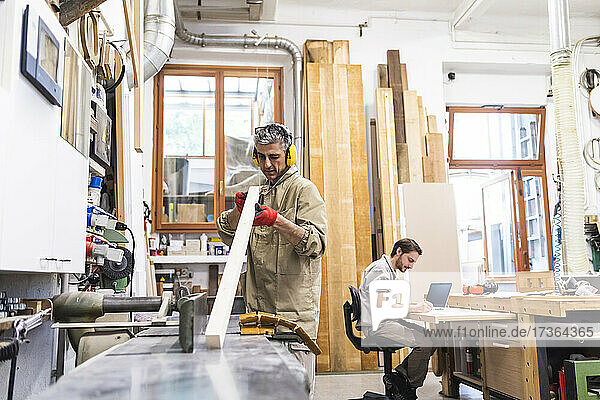 Älterer männlicher Tischler bei der Arbeit an Holz  während ein Kollege in der Werkstatt schreibt