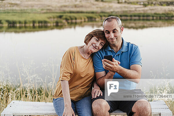 Sohn benutzt sein Smartphone  während er mit seiner Mutter auf einer Bank am See sitzt