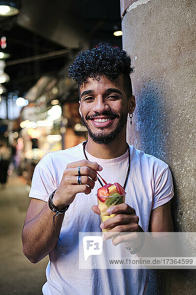 Lächelnder junger Mann mit Obstsalat an der Wand stehend