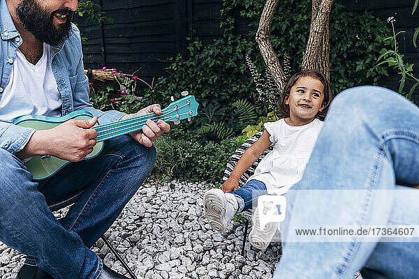Smiling girl looking at mother while father playing guitar in back yard