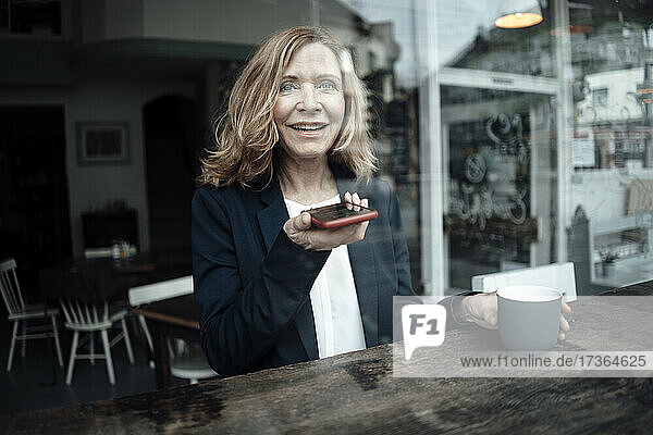 Lächelnde ältere Geschäftsfrau  die eine Kaffeetasse in der Hand hält  während sie über einen Lautsprecher mit ihrem Smartphone spricht  während sie in einem Café sitzt