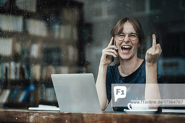 Fröhliche Geschäftsfrau  die mit ihrem Smartphone spricht und den Mittelfinger durch das Fenster eines Cafés zeigt
