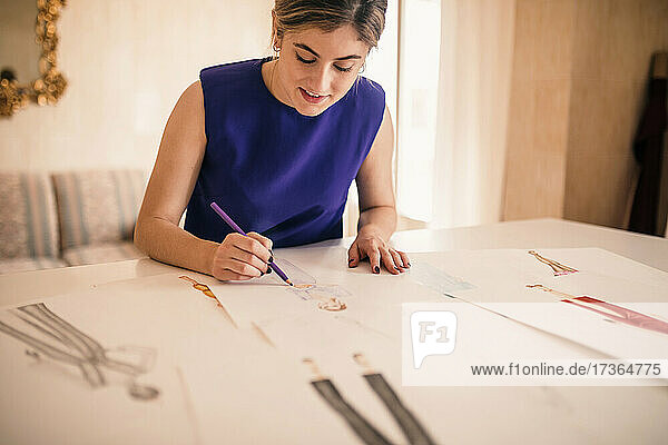 Junge Modeexpertin  die an einer Werkbank sitzend auf Papier zeichnet