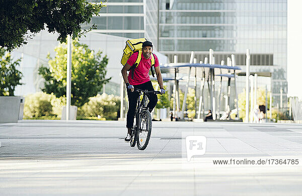 Smiling delivery man wearing yellow backpack riding bicycle in city