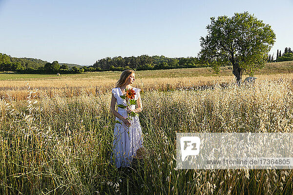 Junge Frau hält Sonnenblume  während sie inmitten von Gras im Feld steht