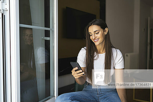 Glückliche schöne Frau  die mit ihrem Handy im Internet surft  während sie am Fenster sitzt