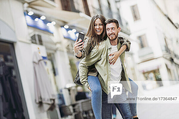 Smiling woman taking selfie through smart phone while piggybacking on boyfriend in city