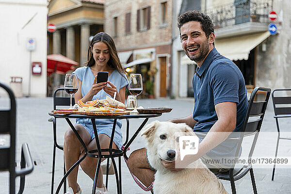 Fröhlicher Mann sitzt mit Hund und Frau in einem Straßencafé