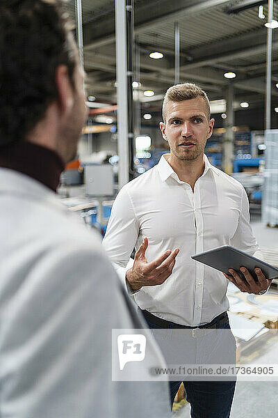 Geschäftsmann hält ein digitales Tablet in der Hand  während er sich mit einem Kollegen in einer Fabrik unterhält
