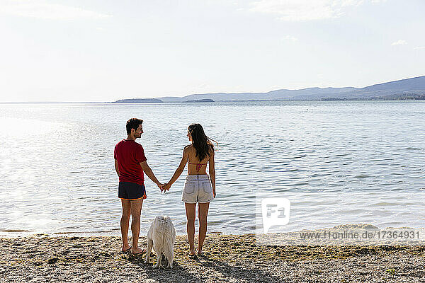 Paar hält sich an den Händen  während es mit seinem Hund am Seeufer steht