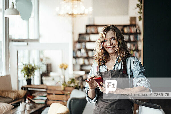 Lächelnde Café-Besitzerin  die ihr Smartphone hält  während sie in einem Café steht
