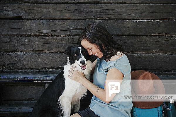 Lächelnde Frau mit geschlossenen Augen  die einen Hund umarmt  während sie vor einer Hütte sitzt