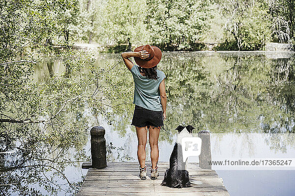 Mittlere erwachsene Frau mit Hut steht mit ihrem Hund auf einem Steg an einem See im Wald