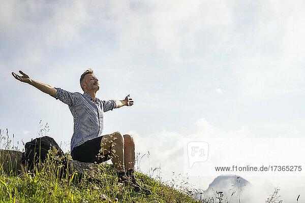 Carefree man with arms outstretched sitting on mountain