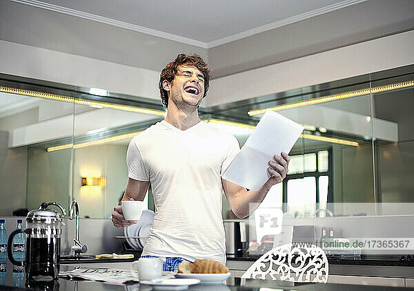 Glücklicher Mann mit Brief  der lachend eine Kaffeetasse in der Küche hält