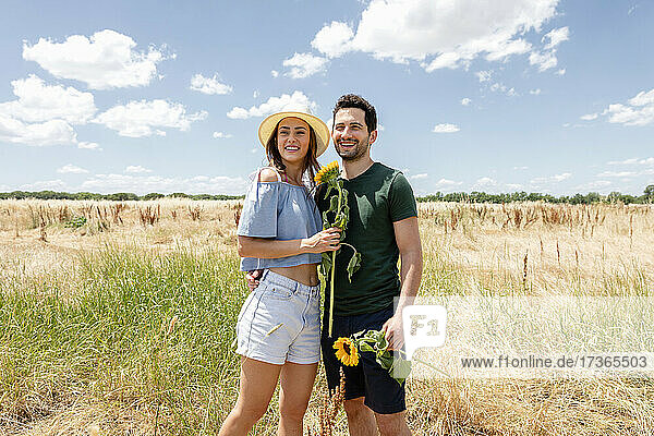 Smiling couple holding sunflowers while standing at meadow