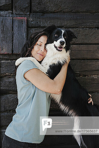 Woman with eyes closed hugging pet dog while standing in front of cottage