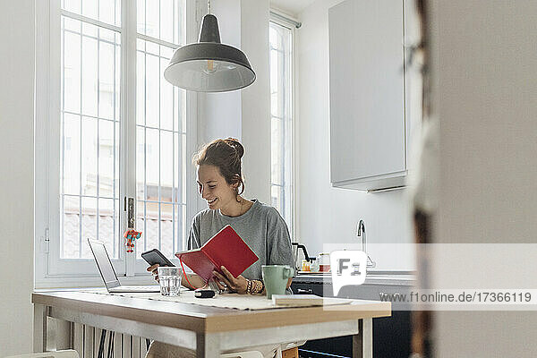 Lächelnde Freiberuflerin  die ein Mobiltelefon benutzt  während sie mit einem Buch in der Küche sitzt