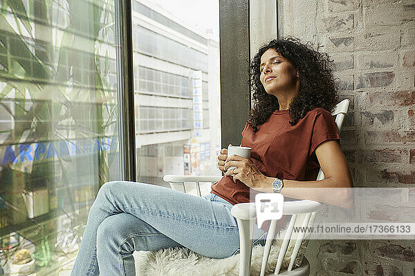 Entspannte Geschäftsfrau  die eine Kaffeetasse hält und mit geschlossenen Augen auf einem Stuhl am Fenster sitzt