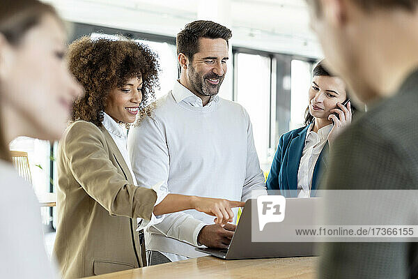 Smiling Afro businesswoman discussing with male professional over laptop while standing at desk in office