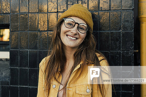 Lächelnde rothaarige Frau mit gelber Jacke  die sich an eine schwarze Wand lehnt