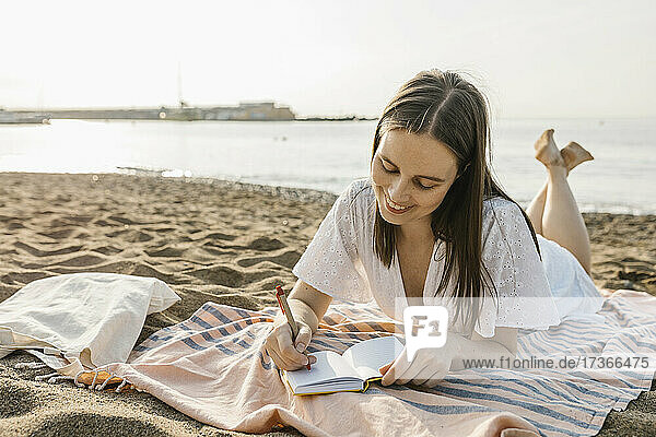 Lächelnde Frau  die in ein Buch schreibt  während sie auf einem Handtuch am Strand liegt