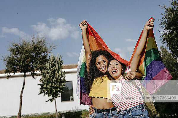 Glückliches junges lesbisches Paar hält Regenbogenflagge unter Himmel