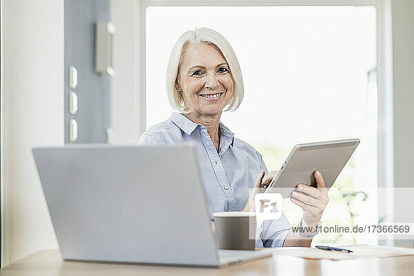 Lächelnde Geschäftsfrau  die ein digitales Tablet am Schreibtisch im Heimbüro hält