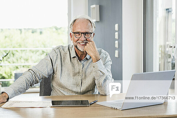 Lächelnder männlicher Berufstätiger mit Hand am Kinn am Schreibtisch im Heimbüro