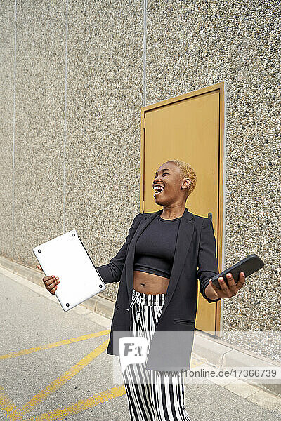Lachende Geschäftsfrau mit Laptop und Mobiltelefon auf dem Fußweg