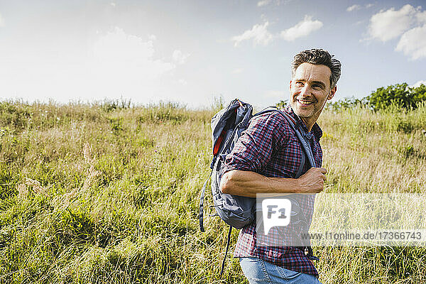 Lächelnder Mann mit Rucksack beim Wandern auf einer Wiese an einem sonnigen Tag