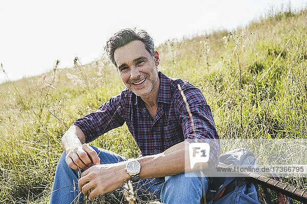 Lächelnder Mann sitzt mit Rucksack und Gitarre im Gras
